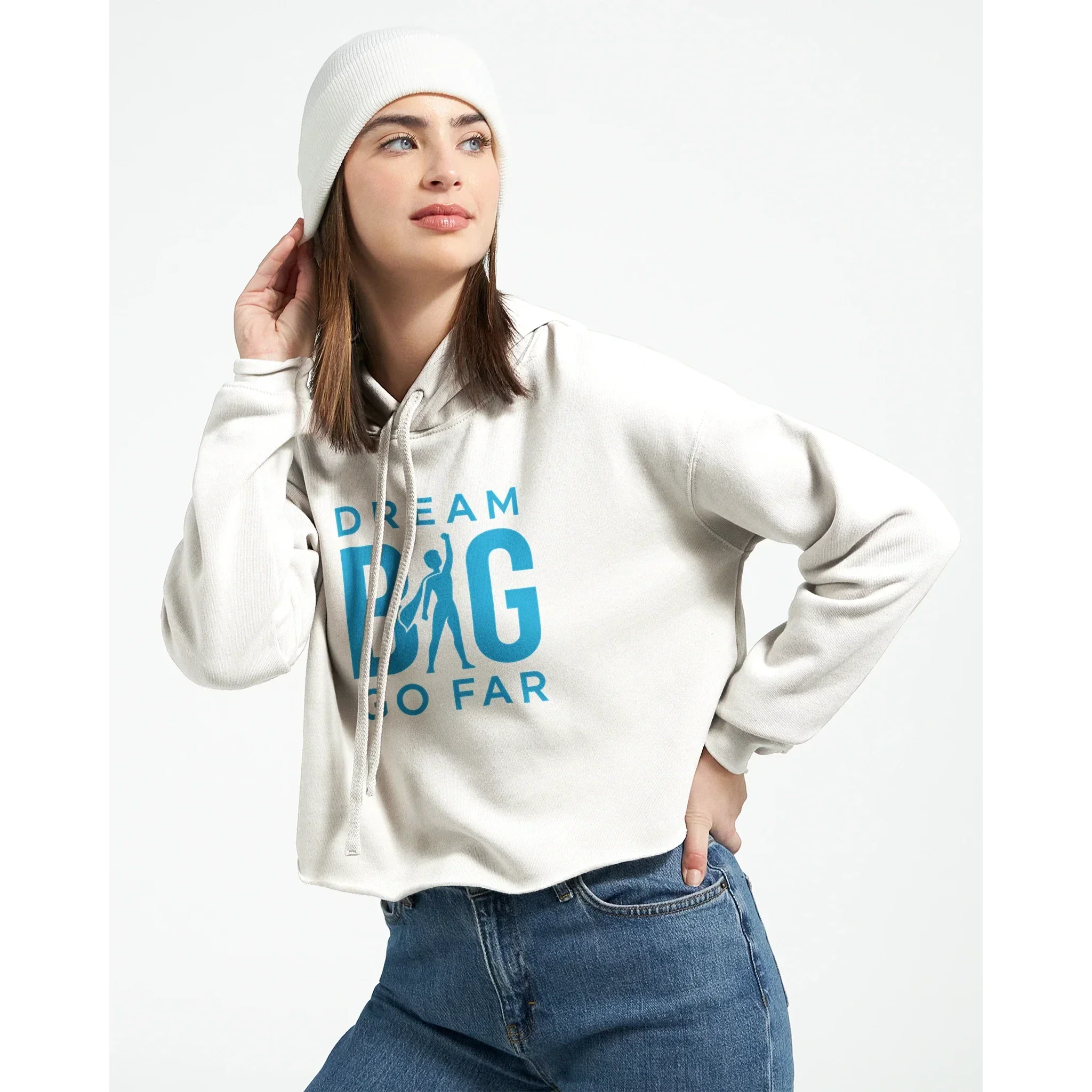 Woman wearing white cropped hoodie with "Dream BIG Go Far" motivational text in blue, paired with a white beanie and blue jeans