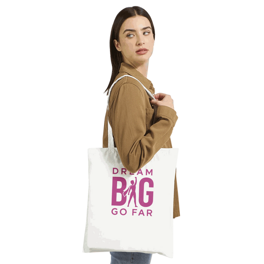 White cotton tote bag with pink "Dream BIG Go Far" motivational print, worn by a woman.